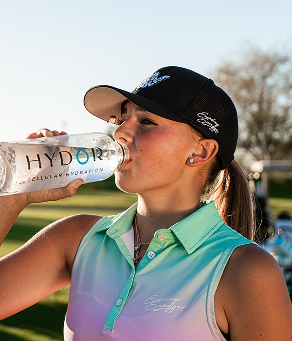 Person drinking a bottle of Hydor cellular hydration water to stay hydrated on a sunny day outdoors.